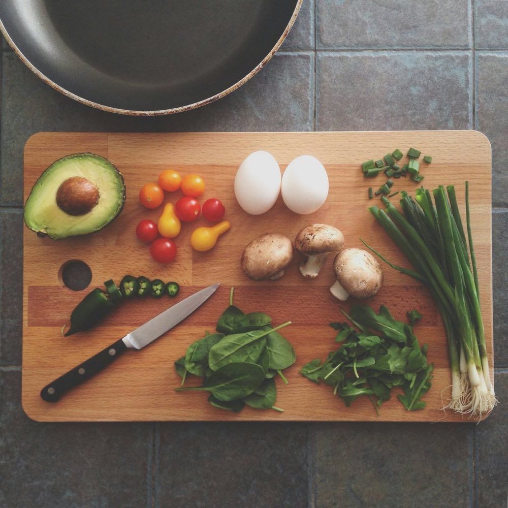 A wooden board with avocado, eggs, mushrooms, tomatoes and greens ready to cook